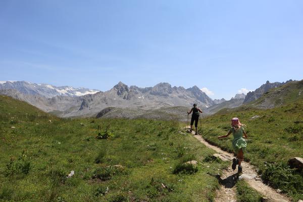 A person with hiking poles walks along a winding dirt path through a vibrant green mountain landscape, while a child in a dress joyfully runs ahead